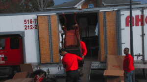 Two men are unloading a parcel from a van. One man is standing inside the back of the van, carefully lifting the parcel and handing it to the other man, who is positioned on the ground.